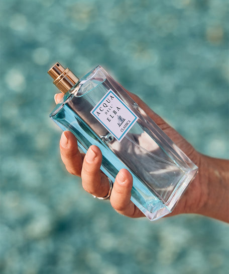 A hand holds a rectangular bottle of Acqua dell'Elba Classica Donna - 100ml by Acqua dell'Elba, filled with light blue liquid, set against a blurred blue water background, capturing the floral and marine essence of the Mediterranean.