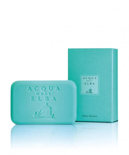 A bar of Acqua dell'Elba Classica Donna Body Soap, in turquoise and embossed with "Acqua dell' Elba," emits a subtle floral scent and sits before its complementary rectangular box. Both the high-end packaging and the soap present a minimalist design adorned with a small ship illustration.