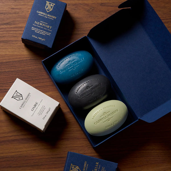 A navy blue Caswell~Massey Classic Collection Soap Set box sits open on wood, revealing three triple-milled, plant-based soaps—teal, black, and cream—alongside nearby "NEWPORT" and "OATMEAL" Caswell-Massey boxes.