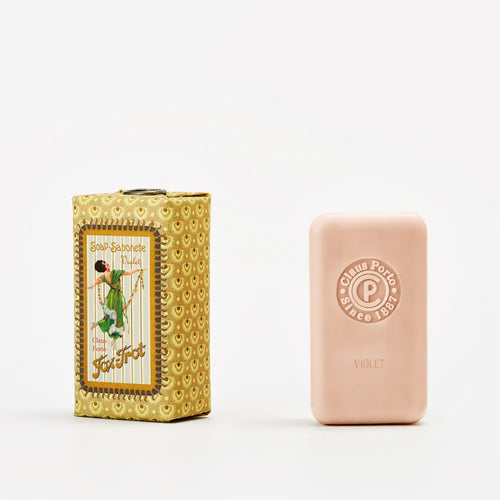 A pink Claus Porto 1887 Classico Fox Trot Violet Soap, featuring a delicate floral scent, stands upright beside its ornate yellow box with vintage artwork and "Soap Savonette Fox Trot" on the front.