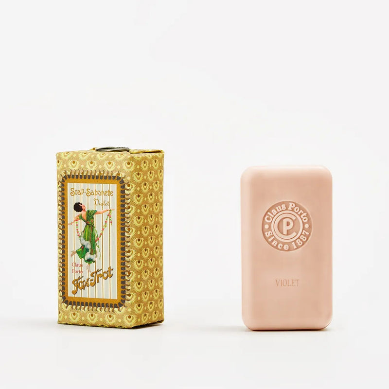 A pink Claus Porto 1887 Classico Fox Trot Violet Soap, featuring a delicate floral scent, stands upright beside its ornate yellow box with vintage artwork and "Soap Savonette Fox Trot" on the front.