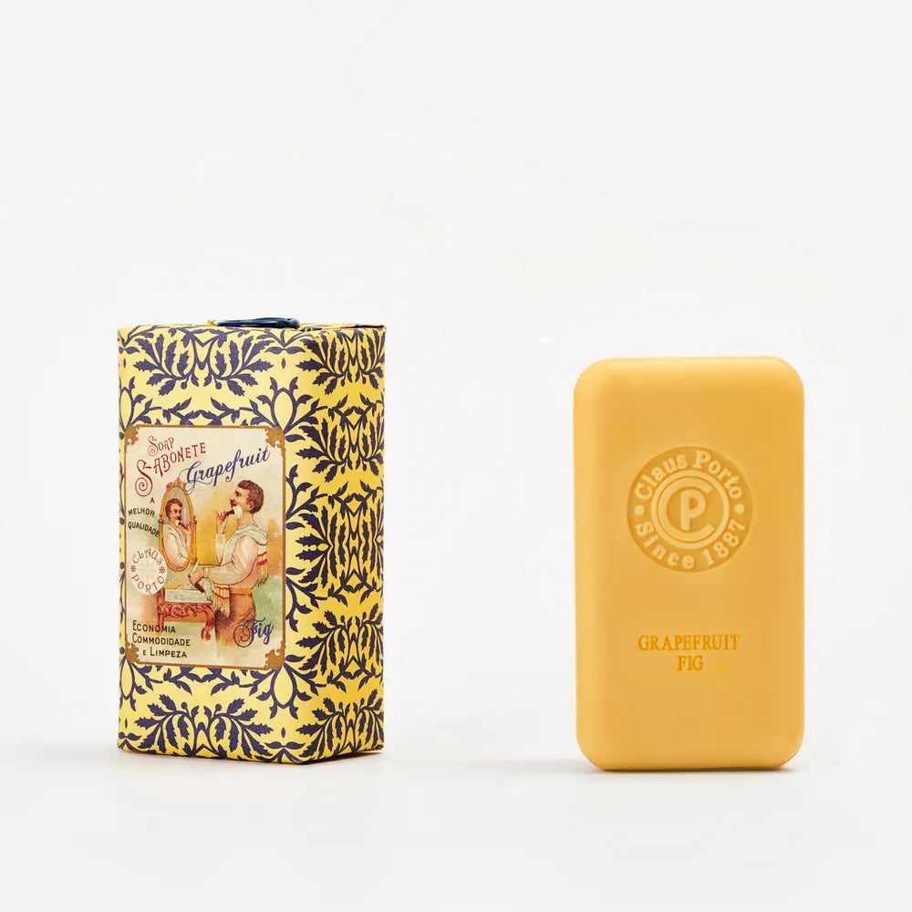 Claus Porto 1887 Classico Barbear Grapefruit Fig Soap is enriched with mango oil. The yellow bar is displayed next to its elegant yellow and blue vintage box, featuring an illustration of two women.
