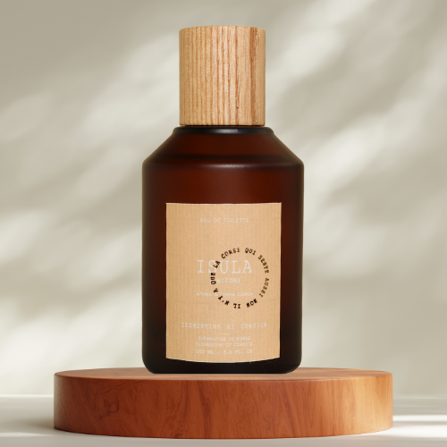 A brown glass ISULA parfums bottle of Clementine eaux de toilette with a wooden cap rests on a round wooden platform. The label shows "ISULA parfums" and "Clementine," evoking a citrus scent. Soft, dappled light falls on a neutral background.