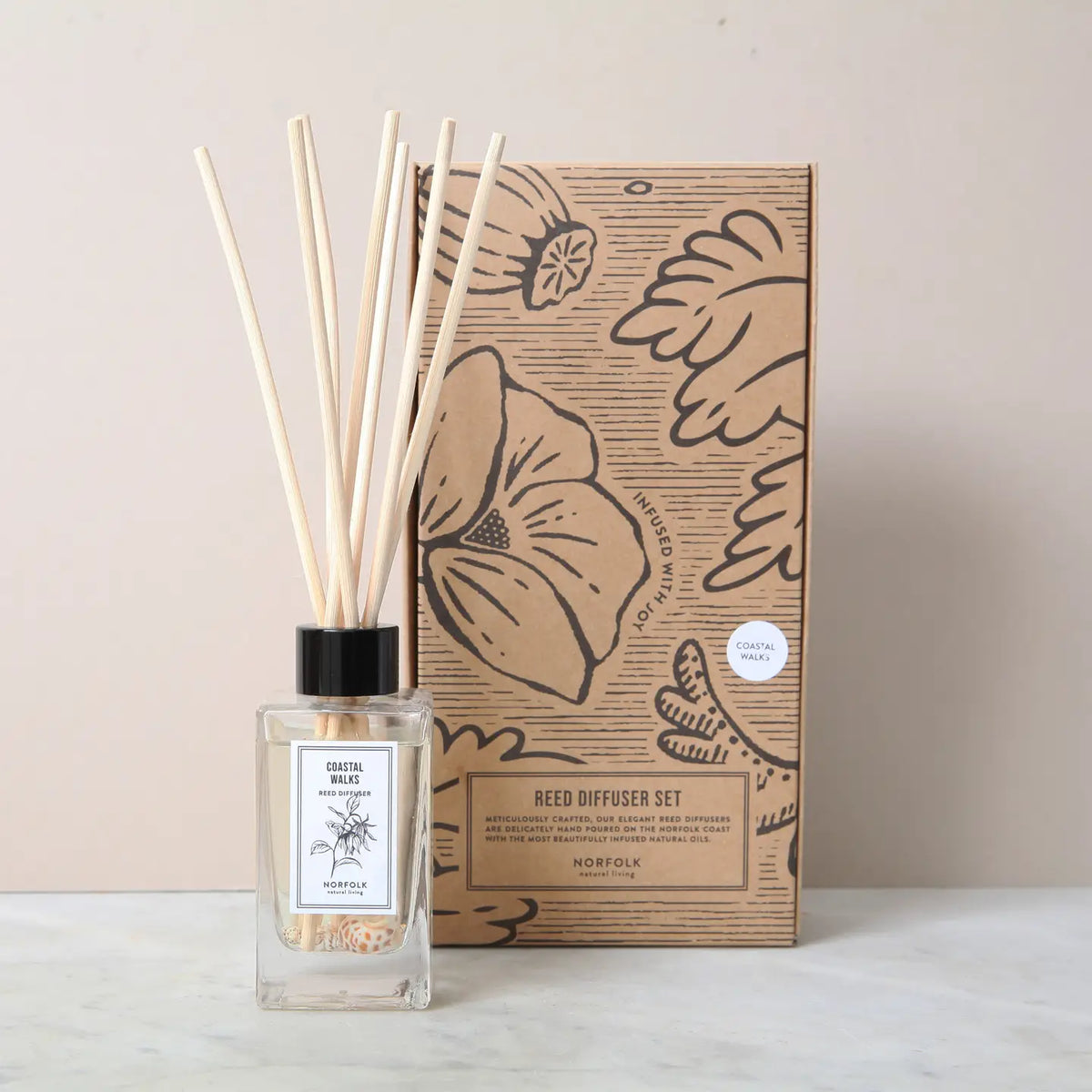 A glass Norfolk Natural Living Coastal Sea Salt Reed Diffuser with wooden sticks stands beside a decorative floral box, offering premium oil that captures the fresh scent of English coast sea salt.