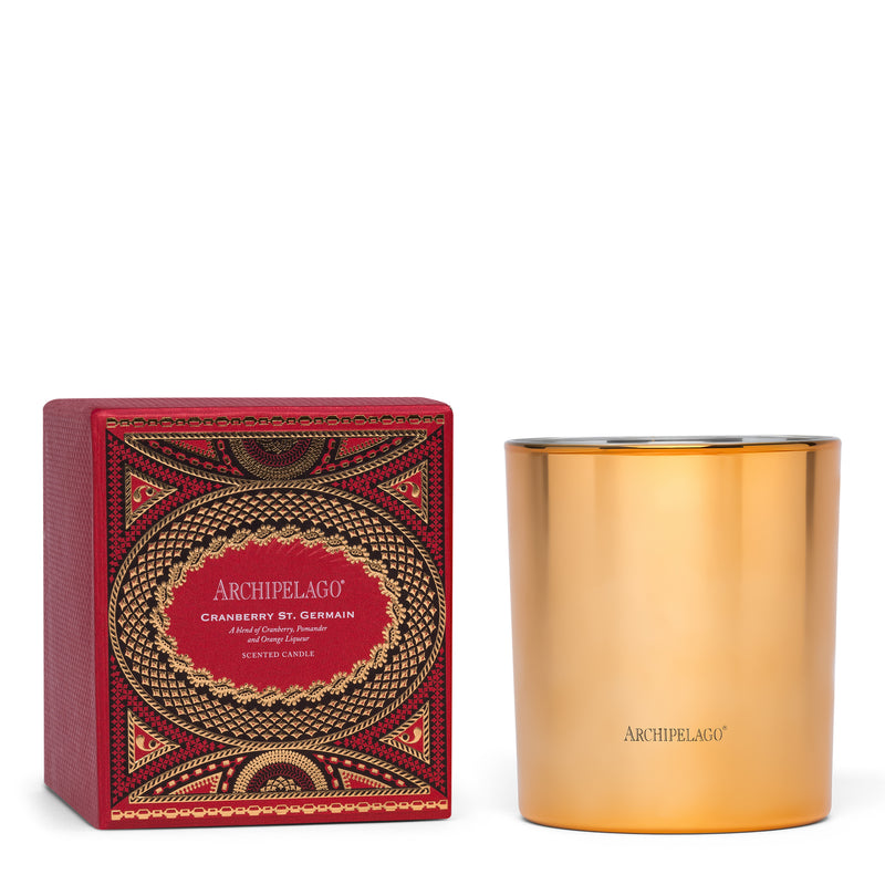 A gold Archipelago Botanicals Cranberry St. Germain Credenza Candle sits beside its festive red and gold box, featuring intricate patterns and elegant labeling that evoke a warm holiday ambiance.
