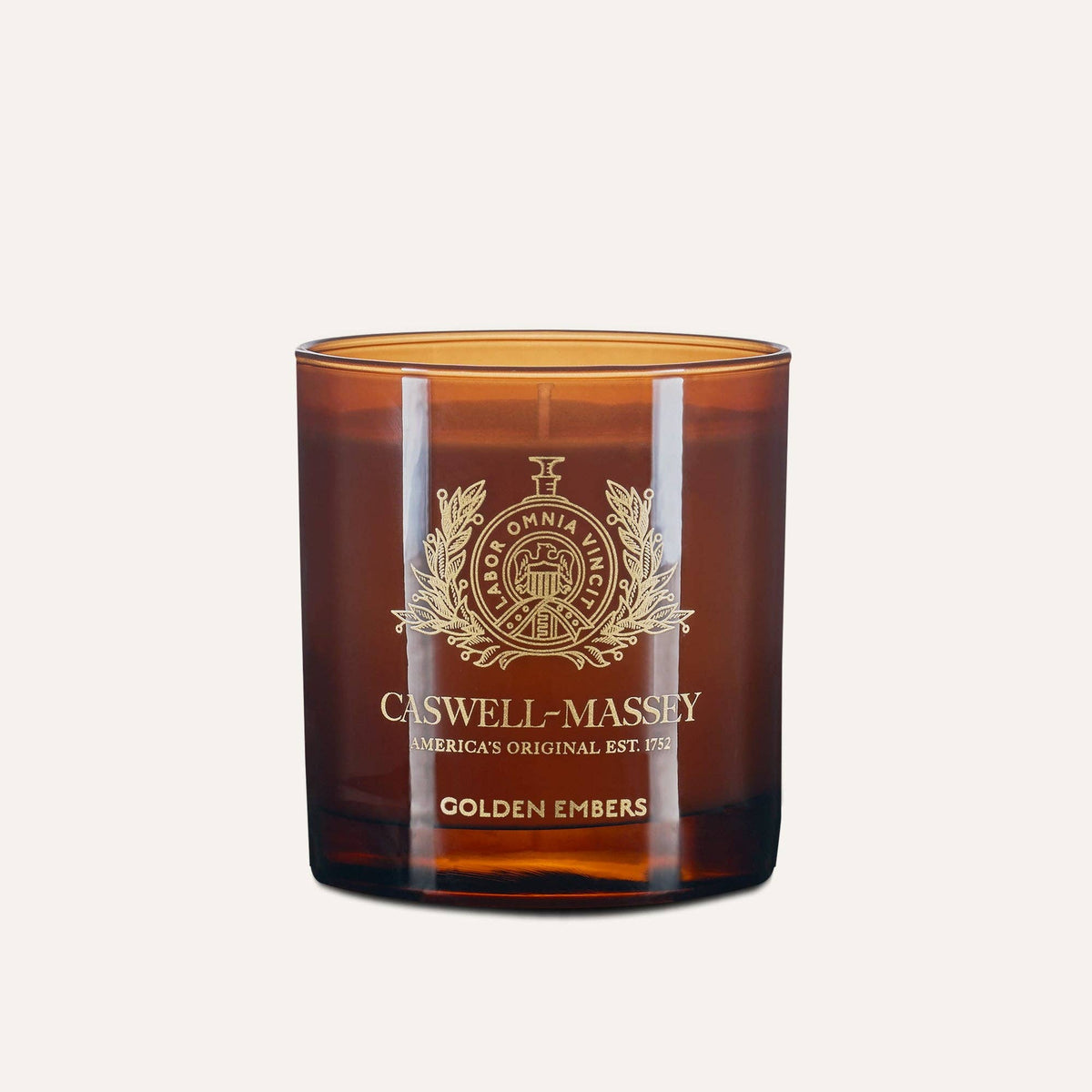 The Caswell-Massey Candle - Golden Embers is a hand-poured soy blend with a meditative scent and stylish gold emblem, showcased against a light beige background.