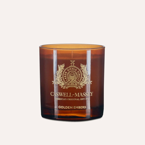 The Caswell-Massey Candle - Golden Embers is a hand-poured soy blend with a meditative scent and stylish gold emblem, showcased against a light beige background.