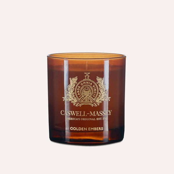 The Caswell-Massey Candle - Golden Embers is a hand-poured soy-blend candle in a brown glass jar with gold embossed branding and "Golden Embers" on the front, featuring a meditative scent against a light background.