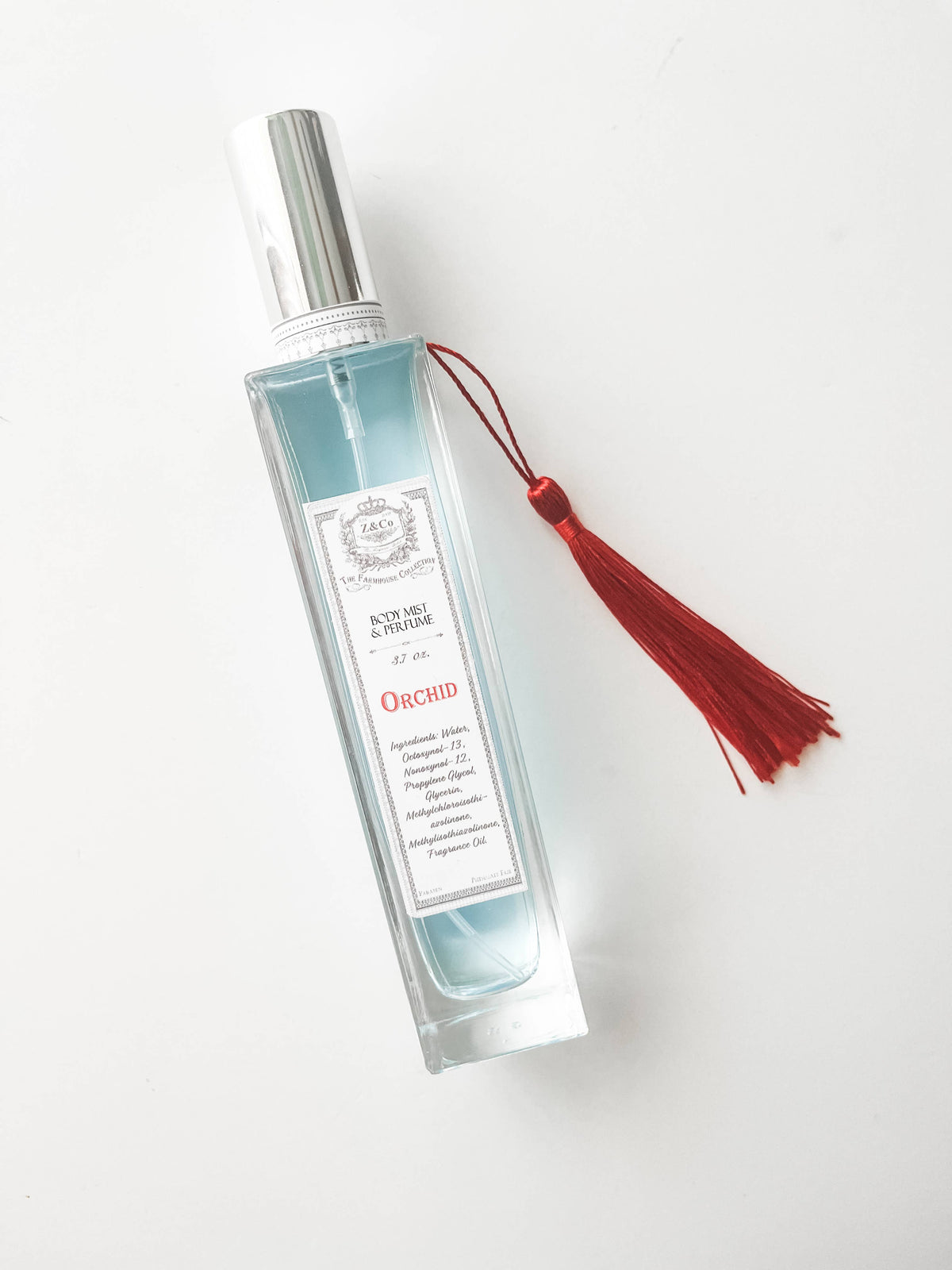 A clear glass bottle with a silver cap, light blue liquid, and a red tassel features the label "Z&Co. Farmhouse Body Mist - Orchid," blending elegance and exotic fragrance by Z&Co. to enhance any room or body.