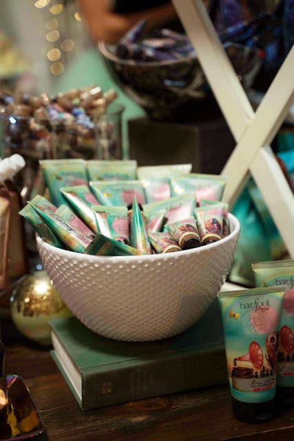 A white textured bowl holds cruelty-free Barefoot Venus Pink Pepper Hand Cream tubes, arranged on stacked green books with more tubes and decor on a wooden table.