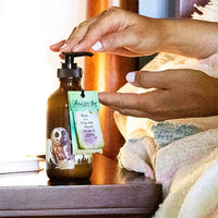 A person dispenses Barefoot Venus Pink Pepper Body Cream from a brown pump bottle with an owl label and note, beside a bed topped with a colorful blanket.