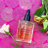 A FRENCH GIRL Organic Hydrating Jasmine Body Oil (2 oz) bottle rests on vibrant pink patterned fabric, accented by a pale pink rose and green leaves—reflecting the essence of this nourishing body oil.