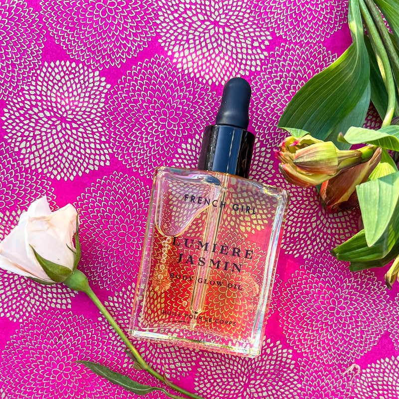 A FRENCH GIRL Organic Hydrating Jasmine Body Oil (2 oz) bottle rests on vibrant pink patterned fabric, accented by a pale pink rose and green leaves—reflecting the essence of this nourishing body oil.