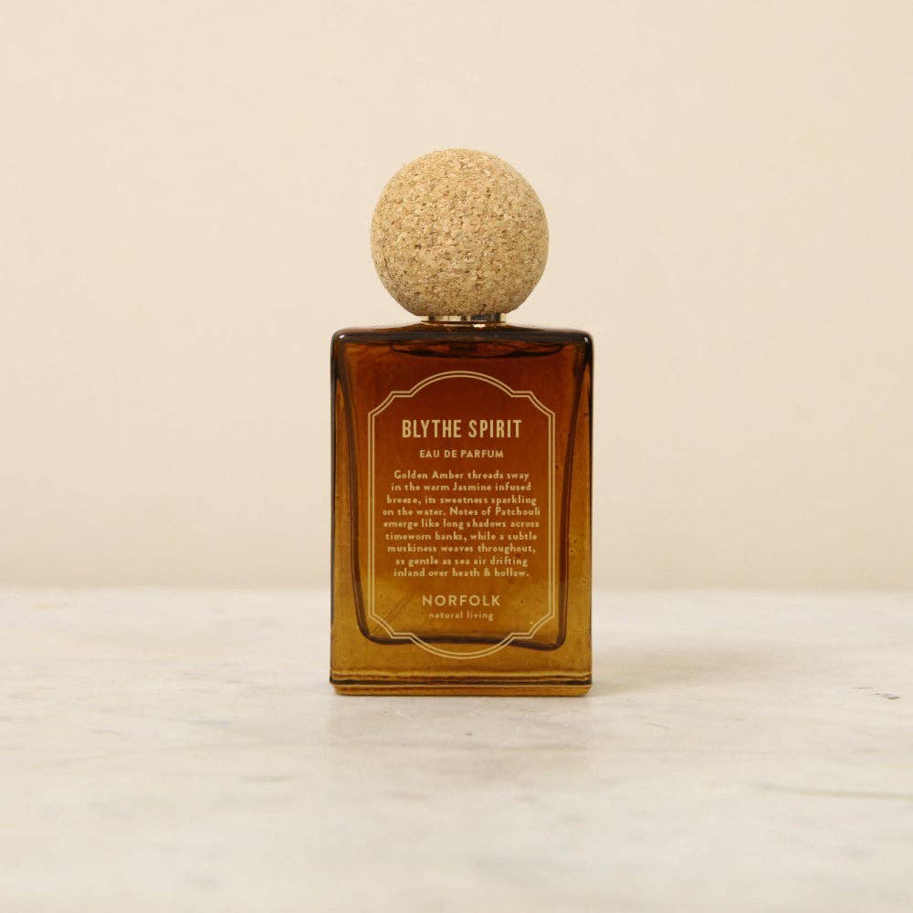 The Norfolk Natural Living Blythe Spirit Eau de Parfum (50ml), in a brown glass bottle with a round cork cap, rests on light marble against a beige background, offering an alluring, long-lasting oil-based fragrance.