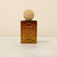 The Norfolk Natural Living Blythe Spirit Eau de Parfum (50ml), in a brown glass bottle with a round cork cap, rests on light marble against a beige background, offering an alluring, long-lasting oil-based fragrance.