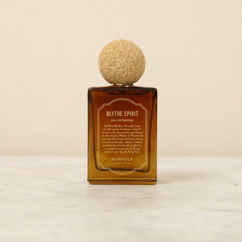 The Norfolk Natural Living Blythe Spirit Eau de Parfum (50ml), in a brown glass bottle with a round cork cap, rests on light marble against a beige background, offering an alluring, long-lasting oil-based fragrance.