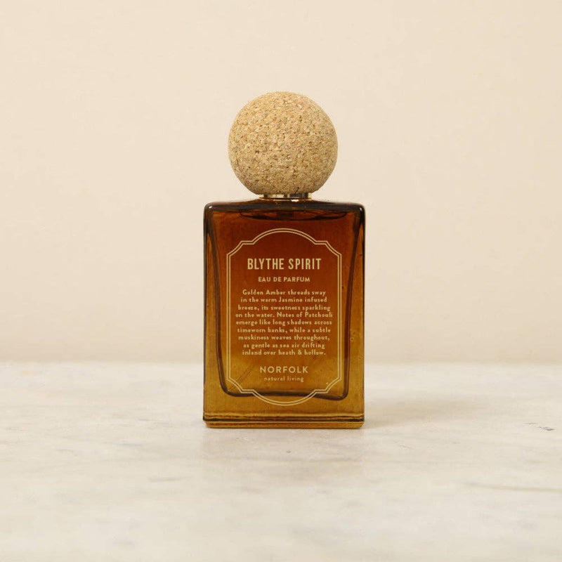 The Norfolk Natural Living Blythe Spirit Eau de Parfum (50ml), in a brown glass bottle with a round cork cap, rests on light marble against a beige background, offering an alluring, long-lasting oil-based fragrance.