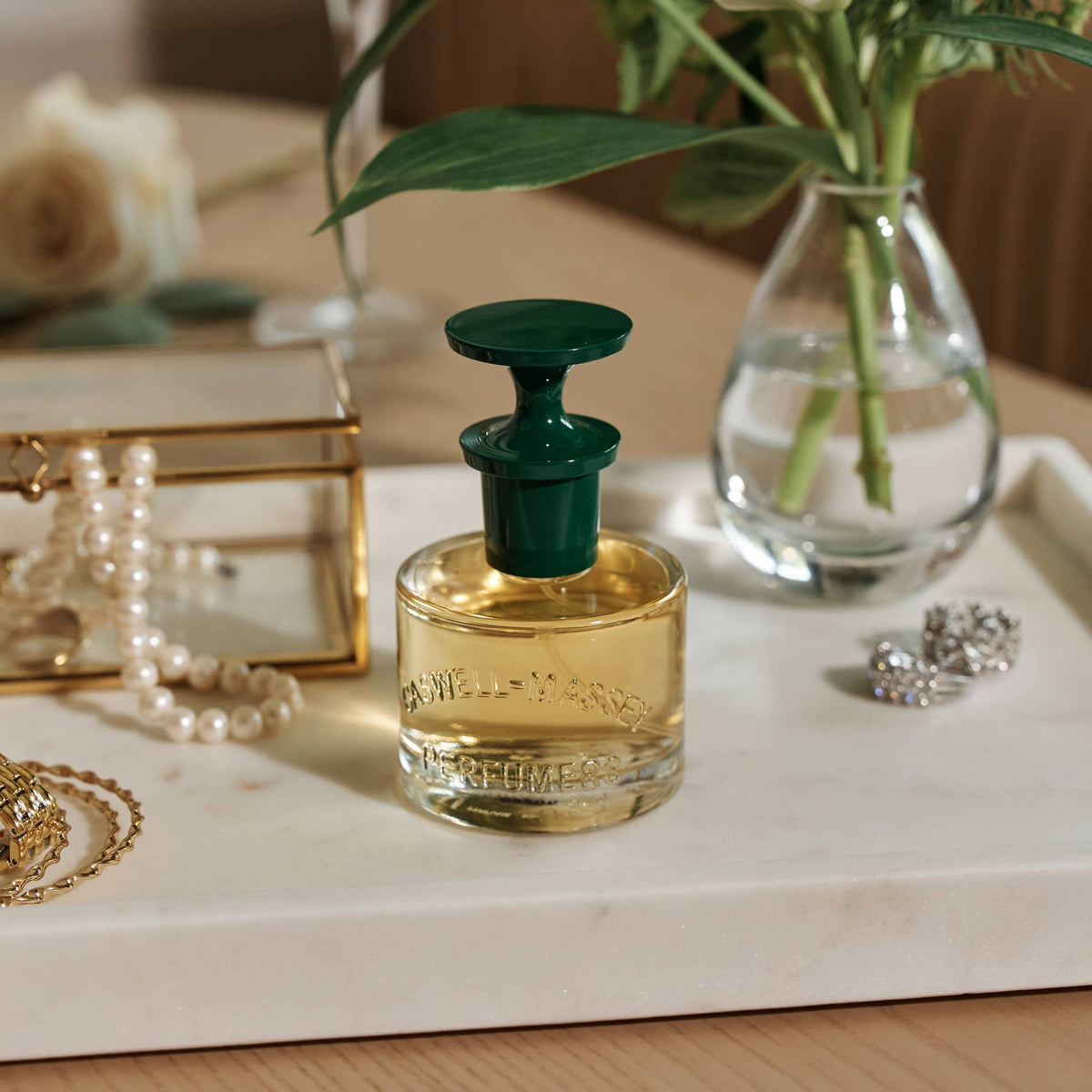 A Caswell-Massey Rose Perfume 60mL bottle with a green cap sits on a white tray, surrounded by jewelry, a pearl necklace, and a vase of green leaves in water atop a wooden surface.