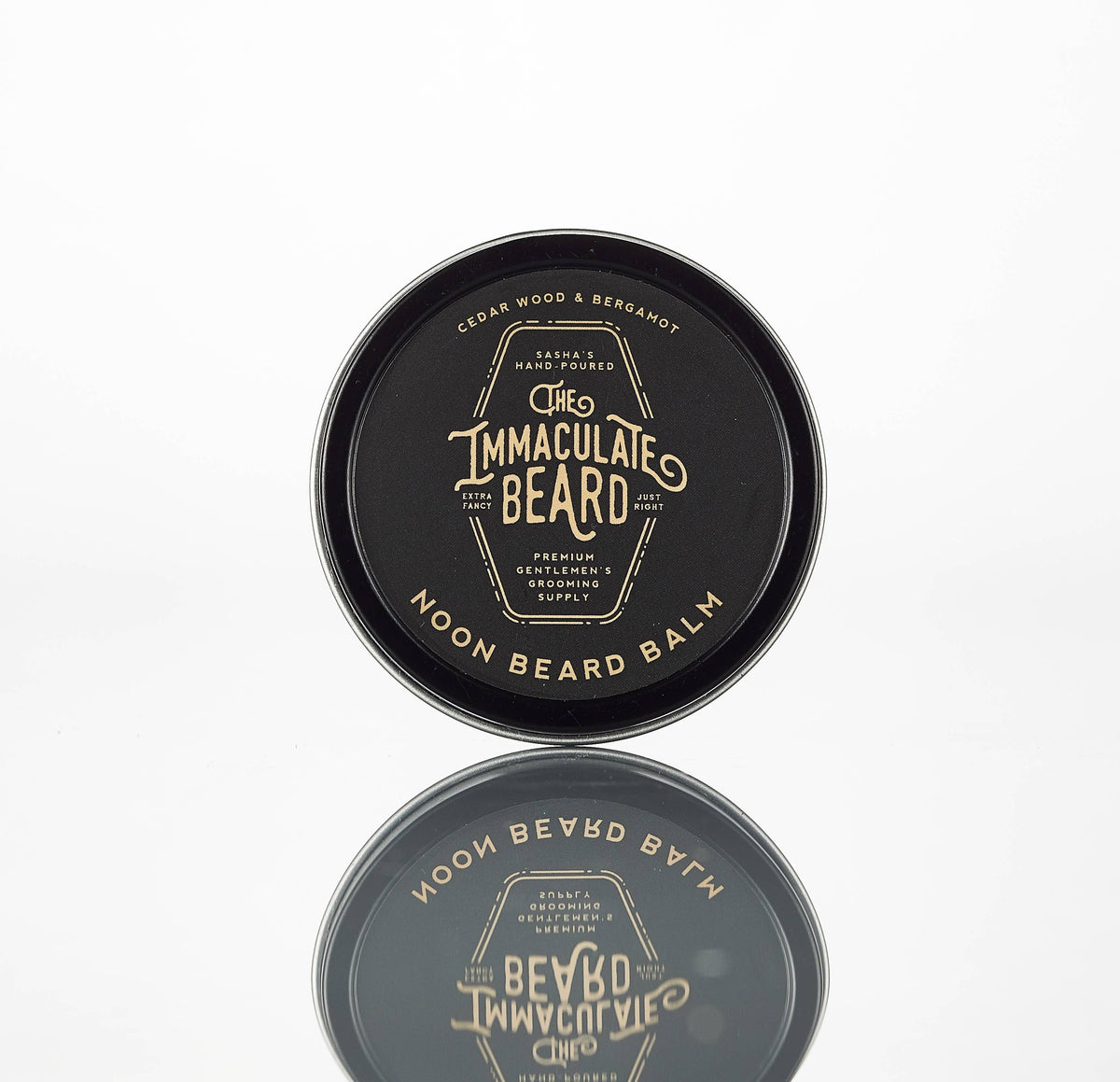 A round black tin labeled "The Immaculate Beard NOON Cologne Oil" with gold text, featuring essential oils—ideal for grooming enthusiasts—reflected on a white surface.