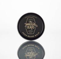 A round black tin labeled "The Immaculate Beard NOON Cologne Oil" with gold text, featuring essential oils—ideal for grooming enthusiasts—reflected on a white surface.