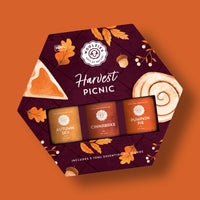 The Woolzies Harvest Picnic Essential Oil Collection features three autumn scents—Autumn Sky, Cinnabake, and Pumpkin Pie—in a hexagonal box with autumn leaves, pie, and a cinnamon roll on an orange background.
