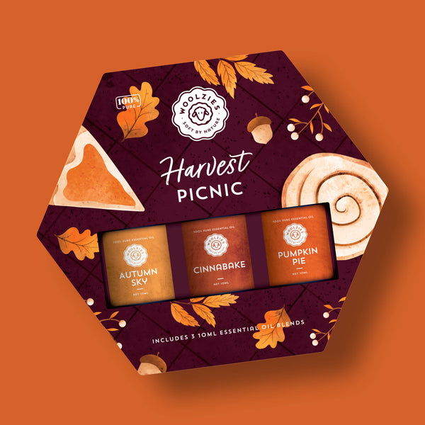 The Woolzies Harvest Picnic Essential Oil Collection features three autumn scents—Autumn Sky, Cinnabake, and Pumpkin Pie—in a hexagonal box with autumn leaves, pie, and a cinnamon roll on an orange background.