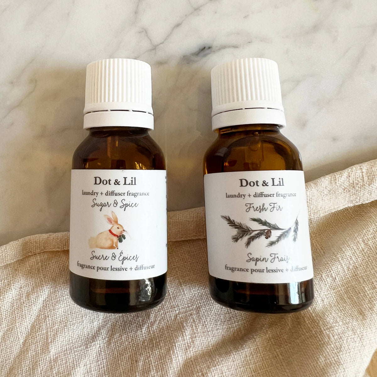 Two brown glass bottles of Dot & Lil laundry + diffuser fragrance sit on a beige cloth. "Sugar & Spice" features a rabbit illustration and holiday notes like ginger cookie; "Fresh Fir" shows a pine branch illustration. Brand: Dot & Lil.