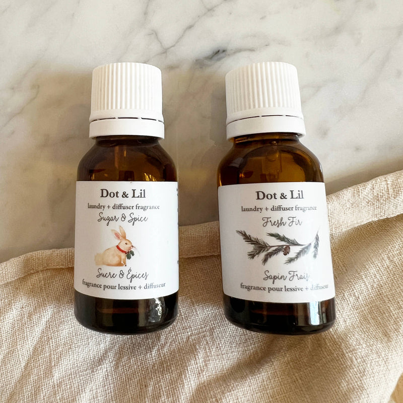 Two brown glass bottles of Dot & Lil laundry + diffuser fragrance sit on a beige cloth. "Sugar & Spice" features a rabbit illustration and holiday notes like ginger cookie; "Fresh Fir" shows a pine branch illustration. Brand: Dot & Lil.