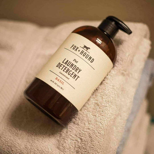 A brown pump bottle of Fox + Hound Pet Natural Laundry Detergent Basil sits on a neatly folded beige towel, emphasizing its refreshing basil scent.