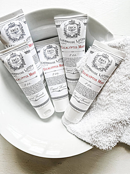 A white dish holds five tubes of Eucalyptus Mint Farmhouse Lotion by Z&Co., perfect travel-sized companions that deeply hydrate your skin. The tubes are white with black and red text. A folded white towel is placed beside the dish, partially overlapping it. The background surface is white.