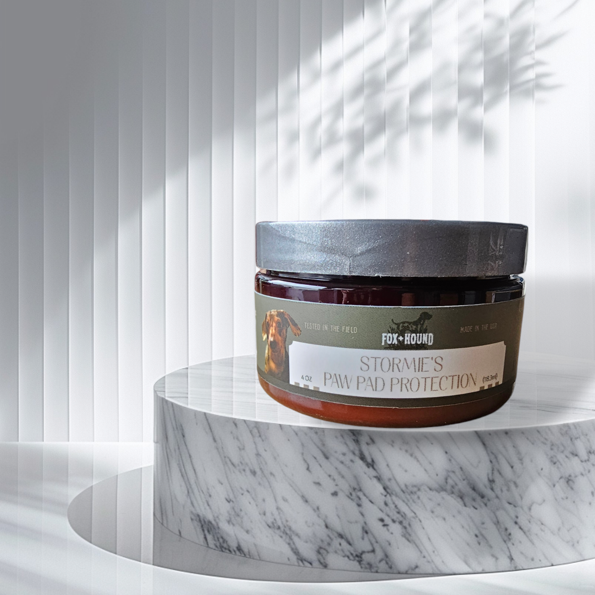 A jar of Fox + Hound Stormies Paw Protection - All Season sits on a round marble pedestal, with vertical white blinds and soft shadows in the background, highlighting this essential dog paw care product.