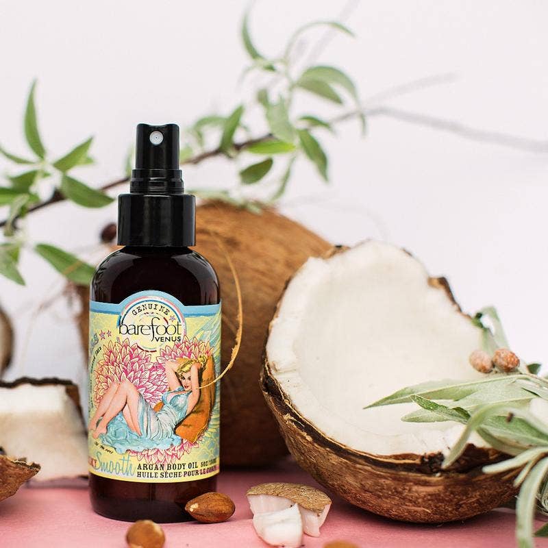 A bottle of Barefoot Venus Coconut Kiss Argan Body Oil, made with all-natural ingredients, is placed amid cracked open coconuts, green foliage, and small nuts. The bottle features vibrant artwork of a woman lounging amid flowers and has a spray nozzle. The scene has a natural, tropical feel.