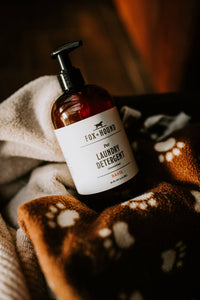 A bottle of Fox + Hound Pet Natural Laundry Detergent Basil rests on a brown blanket with white paw prints, partially covered by a beige towel, filling the air with its fresh basil fragrance.