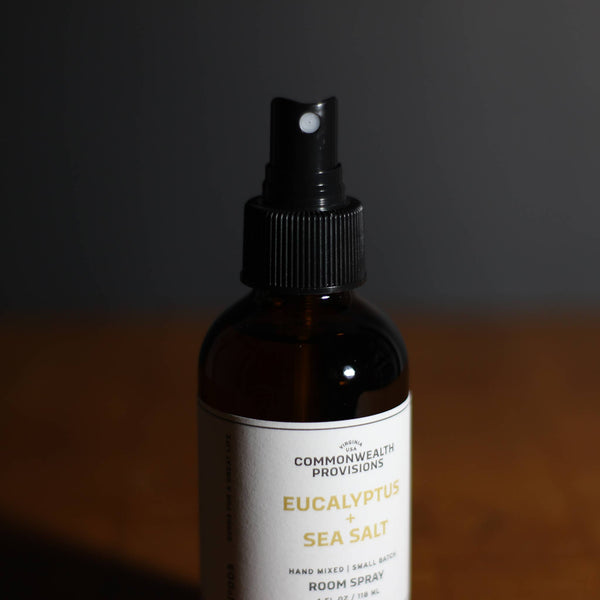The Commonwealth Provisions Eucalyptus + Sea Salt Room Spray sits on a wooden surface, offering fresh eucalyptus and sea salt aromas from its brown bottle, with a dark, blurred background enhancing its calming appeal.