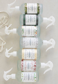Seven Dot & Lil Sugar & Spices Linen + Air Mist bottles are lined up vertically, each with a white spray nozzle and decorative label reflecting scents like Sage, Peppermint, Citrus, and vegan-friendly options.