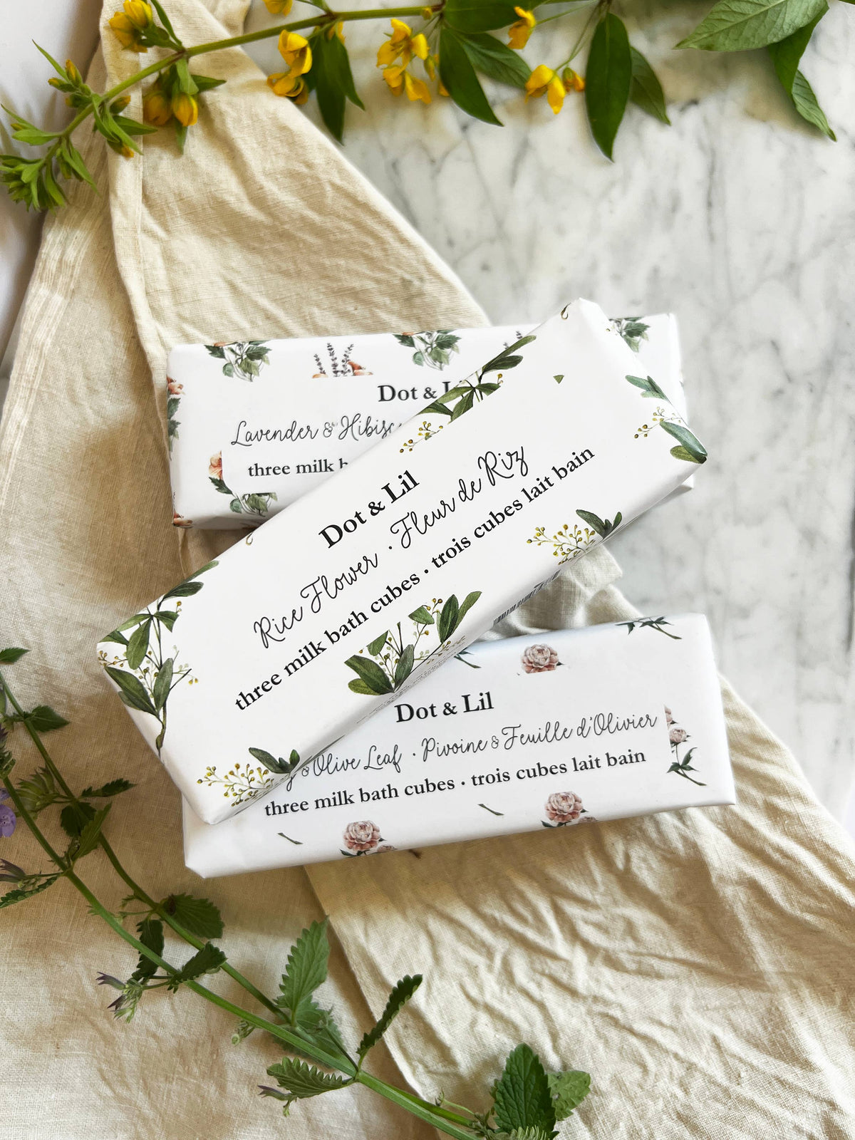 Three floral-wrapped Dot & Lil Lavender & Hibiscus Milk Bath Cubes Trio Gift Sets rest on leaf-patterned fabric with yellow flowers atop a marble surface.