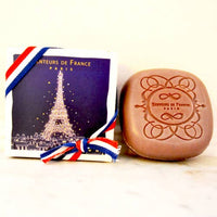 A Senteurs De France Eiffel Tower Amber Scented Soap, embossed with "Senteurs de France Paris" and an ornate border, rests beside a box decorated with the Eiffel Tower and a red, white, and blue ribbon.