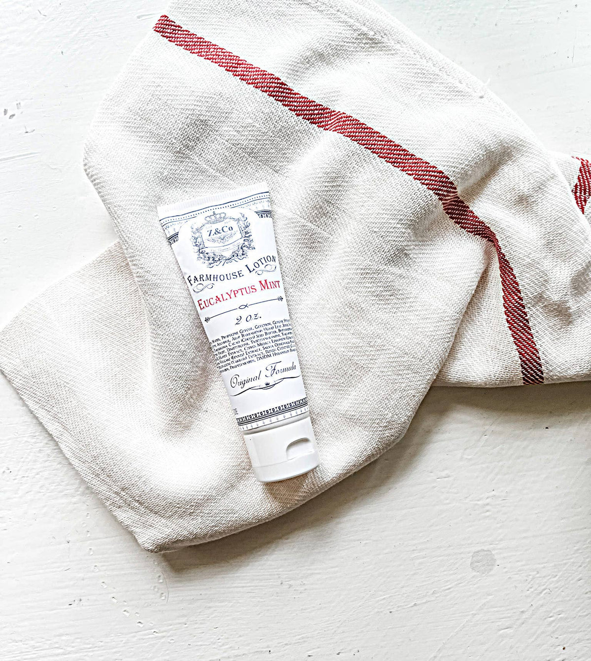 A white and red-striped towel lies on a light-colored surface. On top of the towel, there is a small tube labeled "Z&Co. Eucalyptus Mint Farmhouse Lotion Tube 2 oz," featuring an "Original Formula" text.