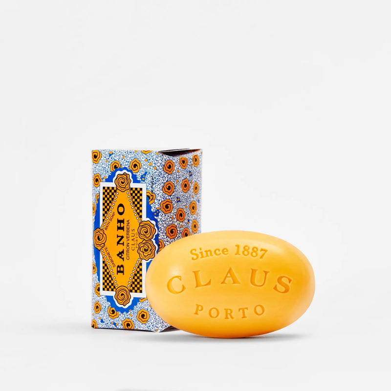 A Claus Porto 1887 Banho Citron Verbena soap bar in bright yellow with embossed lettering, next to its decorative blue and orange patterned packaging, against a white background.