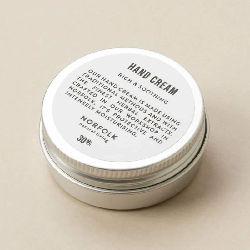 A small, circular container of Norfolk Natural Living Deepest Sleep Nourishing Hand Cream labeled "rich & soothing" with lavender flower oil on a beige background. The jar is made of metal with a detailed text description on the top.