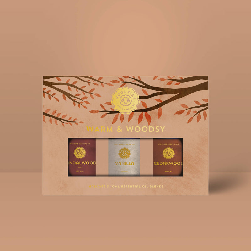 The Woolzies Warm & Woodsy Collection by Woolzies includes three 10ml bottles—sandalwood, vanilla, and cedarwood essential oils—in a rectangular box with autumn leaves in earthy brown and orange tones.
