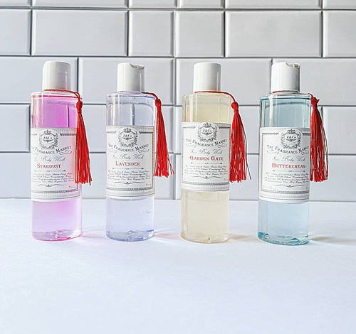 Four bottles of Z&Co. Body Wash Farmhouse Collection displayed against a white tiled background feature signature scents: Stardust (pink), Lavender (purple), Garden Gate (light yellow), and Buttercream (blue). Each bottle is paraben-free and adorned with red tassels.