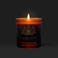 The Caswell-Massey Candle - Cognac & Cashmere glows in a dark amber holder with a gold crest and label, offering an elegant, luxurious scent to any space.