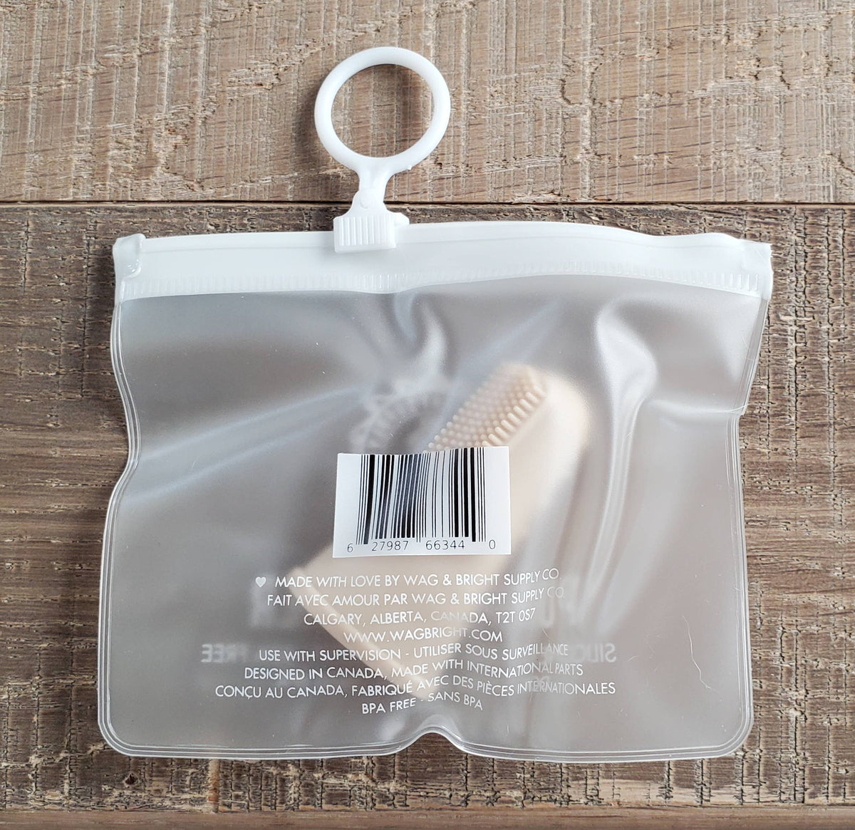 A small transparent pouch featuring a white zip closure and circular hook sits on a wooden surface. Inside, the Wag & Bright Supply Co. - Puppy Polisher Finger Brush with silicone bristles is partially visible through the plastic. The pouch displays a barcode and product information text.