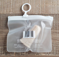 A small transparent pouch featuring a white zip closure and circular hook sits on a wooden surface. Inside, the Wag & Bright Supply Co. - Puppy Polisher Finger Brush with silicone bristles is partially visible through the plastic. The pouch displays a barcode and product information text.