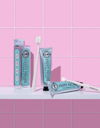 Tubes of toothpaste and BPA-free toothbrushes sit on a white shelf against a pink tile backdrop. The light turquoise packaging with vintage-style labels offers a retro feel, while the Wag & Bright Supply Co.'s Puppy Polisher Pearl Eco Toothbrush in extra small is ideal for keeping your pet's teeth healthy.