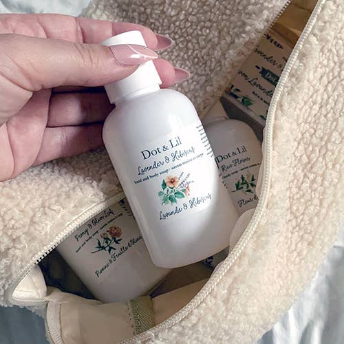 A manicured hand holds a travel-size Dot & Lil Small Lavender & Hibiscus liquid hand + body soap above an open beige toiletry bag filled with similar Dot & Lil products.