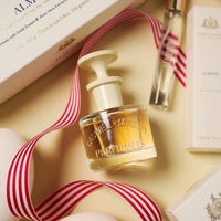 A 60mL bottle of Caswell-Massey Almond Eau de Toilette with a cream cap rests on a beige surface, surrounded by red striped ribbon, boxes, and a small vial, showcasing this floral amber gourmand scent from Caswell-Massey.