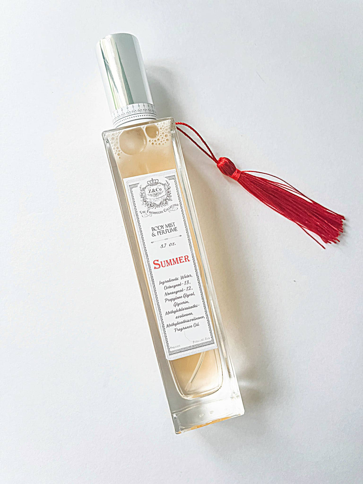 A rectangular glass bottle from Z&Co. Farmhouse Collection Body Mist Perfumes features a silver cap, red tassel, and elegant "Summer" label—perfect for holding your favorite Z&Co. long-lasting scent or body mist.