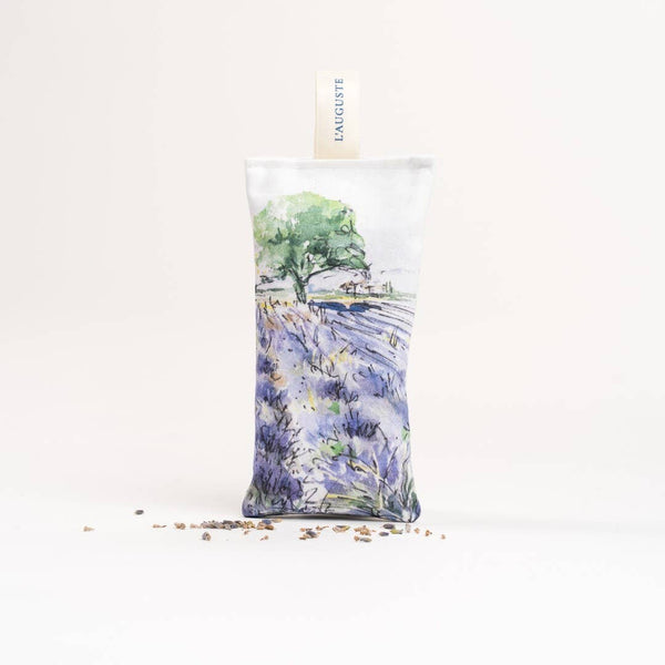 The L'AUGUSTE Provence sachet, handcrafted in Provence, features a painted landscape with a tree, house, and vibrant purple flowers. Standing upright on a light background with scattered lavender pieces, it highlights the benefits of its organic French lavender content.